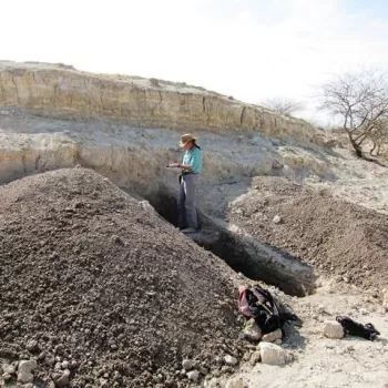 一个女人站在一条地质沟里，面朝灰色的山坡写着笔记。在她的两侧，有两个从战壕里挖出来的棕色土堆